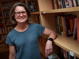 Jocelyn Olcott smiles at the camera. She is standing next to a bookshelf, with her elbow resting on one of the shelves.
