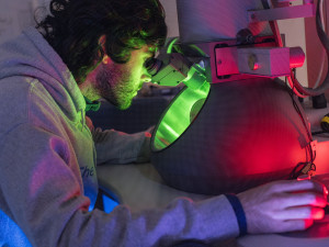Man looks into the lens of a microscope. A green glow emanates from the microscope's body.