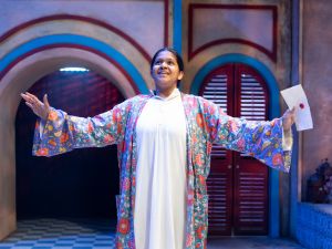 Pillamarri, in a white gown and colorful robe, arms outstretched to the sides, performs in a play. 