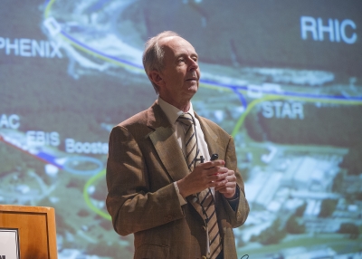 Berndt Mueller stands next to a podium in front of a picture of the Relativistic Heavy Ion Collider